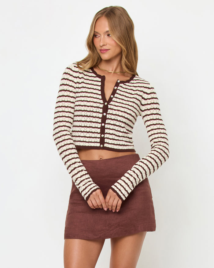 Front view of a model wearing the LSPACE Rosalie Cardigan in cafe-cream, a cropped knit cardigan with cream and dark brown horizontal stripes, V-neckline, and gold button closure, styled with a brown mini skirt.