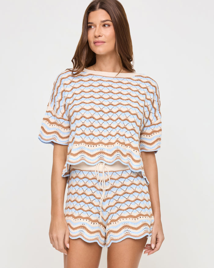 Angled front view of a model wearing the LSPACE Sunchaser Short in north shores, showing the drawstring detail on the waistband and the wave scallop crochet pattern in light blue and tan, paired with the matching top.
