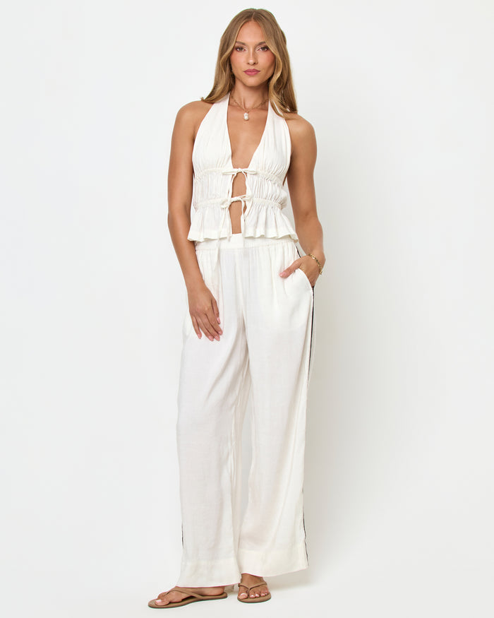 Front view of a model wearing the LSPACE August Top in cream on a white background, showing the plunging V-neckline, double tie-front detail, smocked body, and peplum hem, styled with cream wide-leg pants.