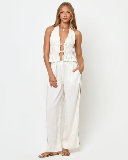 Front view of a model wearing the LSPACE August Top in cream on a white background, showing the plunging V-neckline, double tie-front detail, smocked body, and peplum hem, styled with cream wide-leg pants.