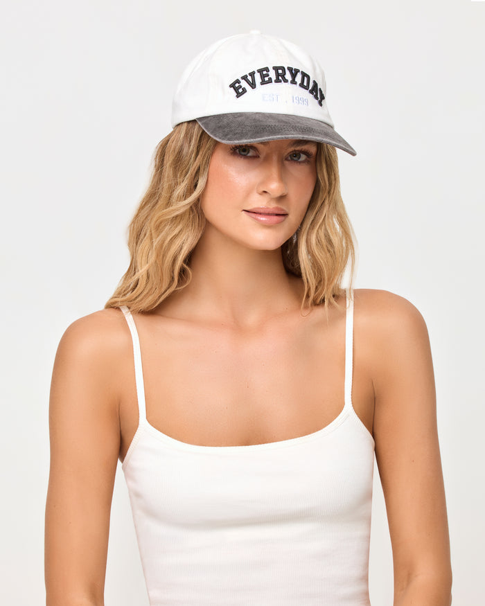 A woman wearing a white baseball cap with a gray brim and 'EVERYDAY' printed in black on the front, paired with a white spaghetti-strap camisole top, against a light gray studio background.
