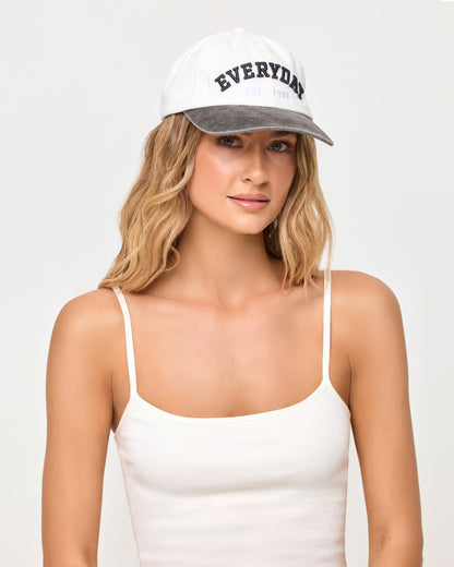 A woman wearing a white baseball cap with a gray brim and 'EVERYDAY' printed in black on the front, paired with a white spaghetti-strap camisole top, against a light gray studio background.