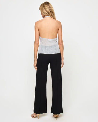 Back view of a model wearing the LSPACE Callie Halter in metallic silver, showing the open back, halter neck tie, and metallic ribbed knit fabric, styled with black wide-leg flare trousers.
