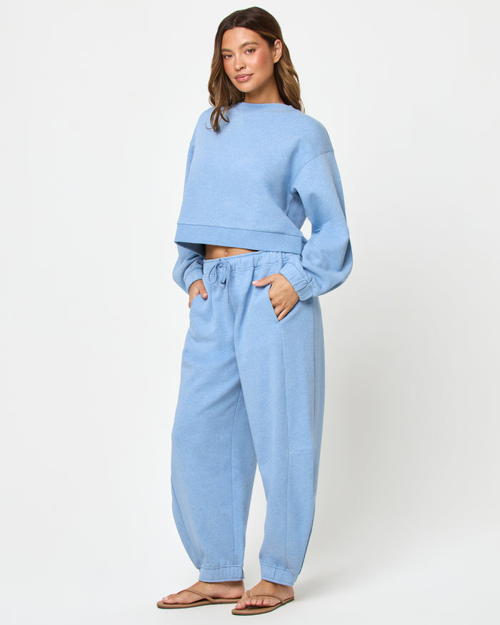 Three-quarter front studio view of the Fleur Sweatshirt and Fleur Pant in chambray blue, a chambray blue cropped crewneck sweatshirt paired with matching chambray blue wide-leg cuffed jogger pants and tan flip flops.