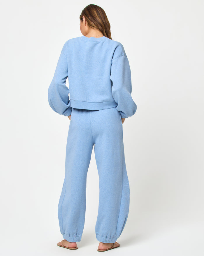 Back full-length studio view of the Fleur Sweatshirt and Fleur Pant in chambray blue, a chambray blue cropped crewneck sweatshirt paired with matching chambray blue wide-leg jogger pants and tan flip flops.