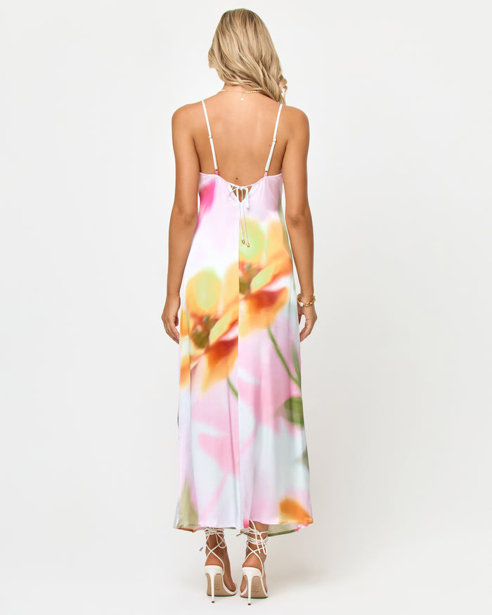 Back view of a model wearing the LSPACE Gabby Maxi in soft focus, highlighting the strappy back tie detail and the soft-focus floral print in yellows, oranges, and pinks on the satin maxi dress.