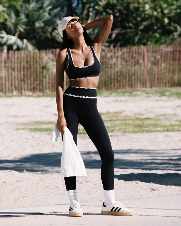 Outdoor lifestyle front full-length view of the Heartbreaker Legging in black, black high-waist leggings with white zig-zag diamond trim at the waist, styled with a matching black sports bra, white baseball cap, white Adidas sneakers with white crew socks, holding a white jacket, against a beach and palm tree background.