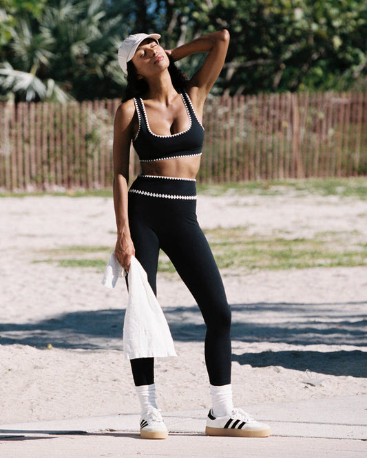 Outdoor lifestyle front full-length view of the Heartbreaker Legging in black, black high-waist leggings with white zig-zag diamond trim at the waist, styled with a matching black sports bra, white baseball cap, white Adidas sneakers with white crew socks, holding a white jacket, against a beach and palm tree background.