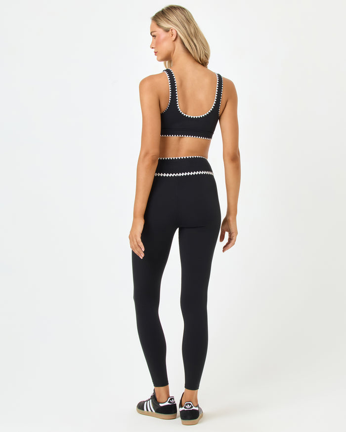 Back full-length studio view of the Heartbreaker Bra and Legging in black, a black sports bra with white zig-zag trim at the back and underbust band paired with matching black high-waist leggings with white zig-zag trim at the waist, styled with Adidas sneakers.
