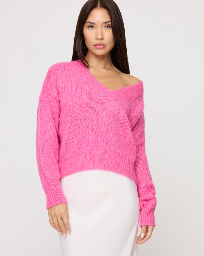 Close-up of a model wearing the LSPACE Icon Pullover - Bubblegum Pink in bubblegum pink, an oversized knit pullover with a wide V-neckline and ribbed hem, styled with a white midi skirt.