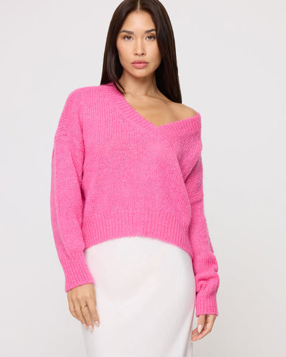 Close-up of a model wearing the LSPACE Icon Pullover - Bubblegum Pink in bubblegum pink, an oversized knit pullover with a wide V-neckline and ribbed hem, styled with a white midi skirt.