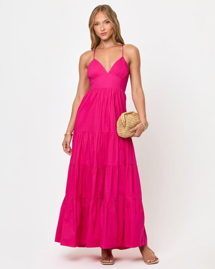 Front view of a model wearing the LSPACE Laina Dress in Bougainvillea on a white background, a hot pink tiered maxi dress with spaghetti straps, V-neckline, fitted bodice, and flowing tiered skirt, styled with a woven rattan clutch.