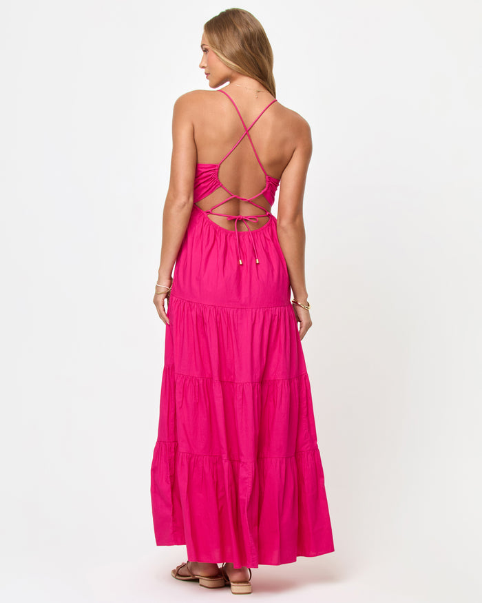 Back view of a model wearing the LSPACE Laina Dress in Bougainvillea on a white background, showing the criss-cross spaghetti straps and lace-up open back detail of the hot pink tiered maxi dress.