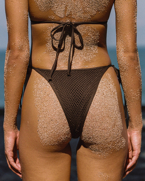 Close-up back view of a model at the beach wearing the LSPACE espresso Levy Bikini Bottom with a crochet mesh texture and side tie detail, with sand-dusted skin visible.