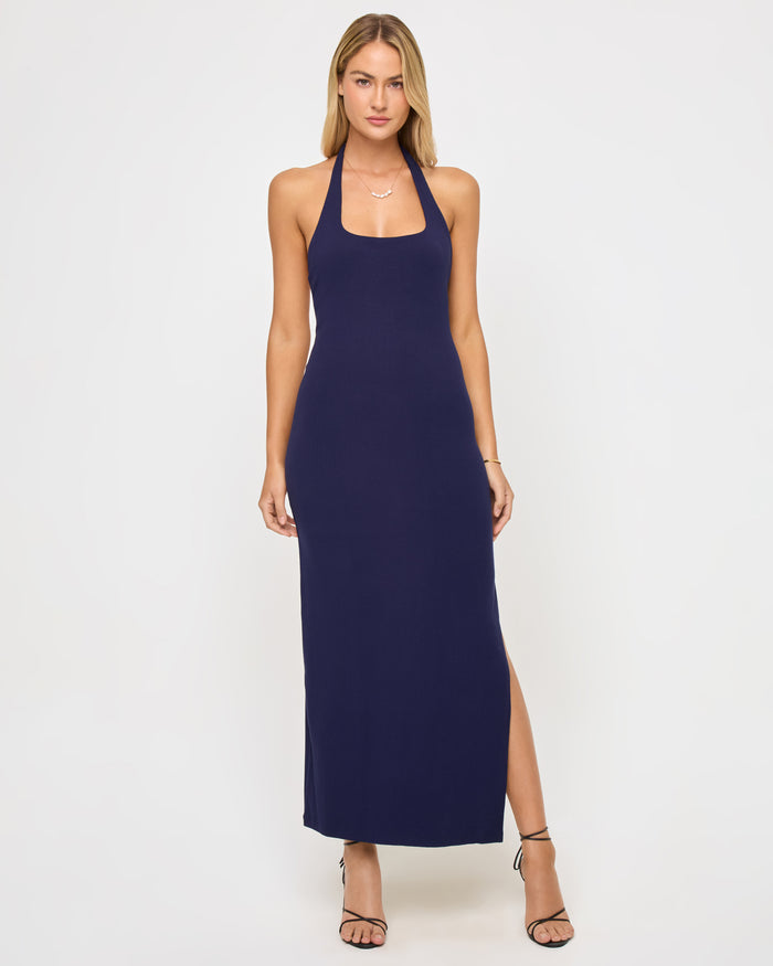 Full-length front view of a model wearing the LSPACE London Dress in midnight blue, a maxi-length halter dress with a square neckline, fitted silhouette, and side slit.
