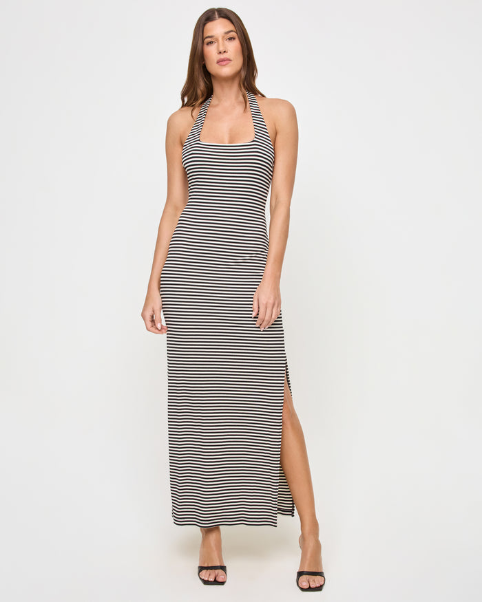 Front view of a model wearing the LSPACE London Dress in see both sides stripe, a fitted maxi dress with a square neckline, halter back strap, horizontal black and cream stripes, and a side slit at the hem.