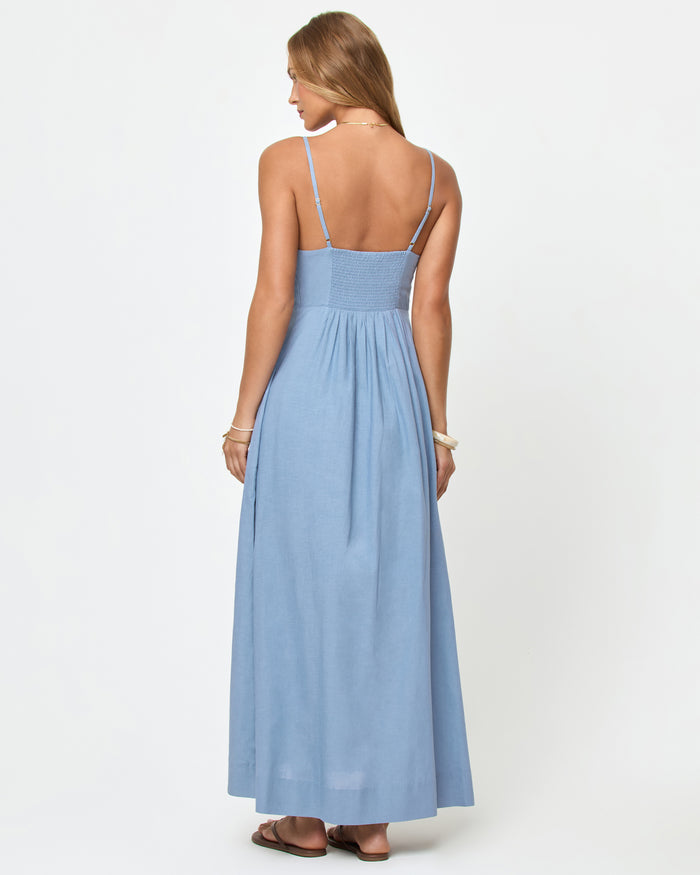 Back view of a model wearing the LSPACE Mabel Maxi Dress in chambray, showing the low open back, spaghetti straps, and smocked back bodice with a flowing A-line skirt.