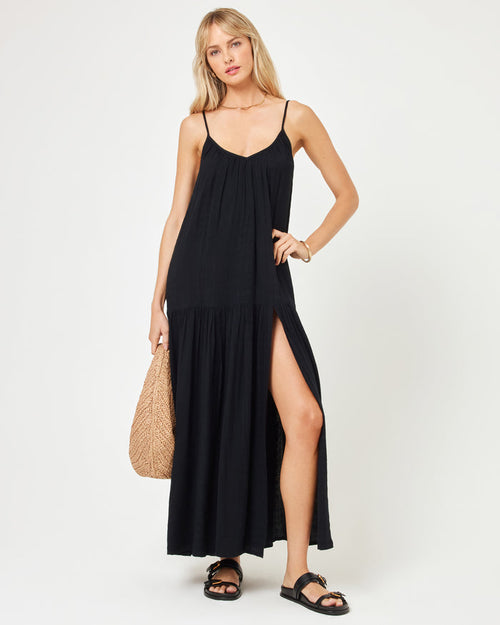 Front view of a model wearing the LSPACE Malibu Dress in black, featuring spaghetti straps, a V-neckline, a flowy tiered skirt with a front slit, and black sandals, holding a woven bag.