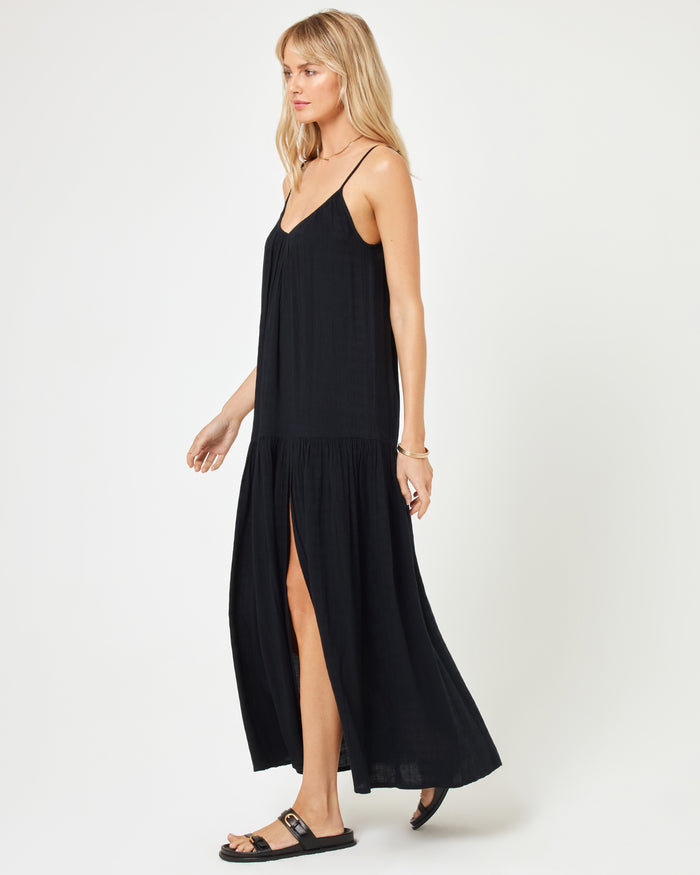 Side view of a model wearing the LSPACE Malibu Dress in black, showcasing the spaghetti straps, tiered skirt, and front slit, paired with black sandals.