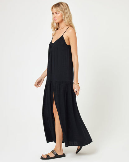 Side view of a model wearing the LSPACE Malibu Dress in black, showcasing the spaghetti straps, tiered skirt, and front slit, paired with black sandals.