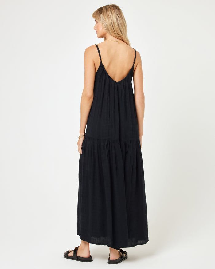 Back view of a model wearing the LSPACE Malibu Dress in black, showing the spaghetti straps, low V-back, and tiered skirt, paired with black sandals.