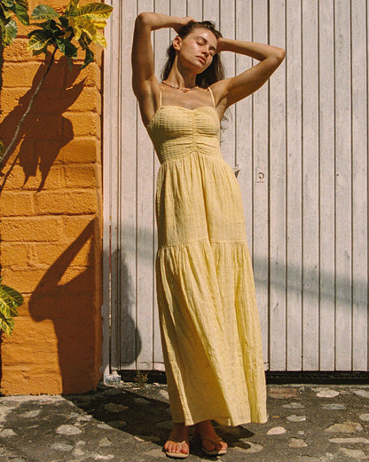 Front view of a model wearing the LSPACE Mallorca Dress in daisy, a yellow maxi dress with a smocked bodice, thin spaghetti straps, and a tiered skirt, photographed outdoors in natural sunlight.