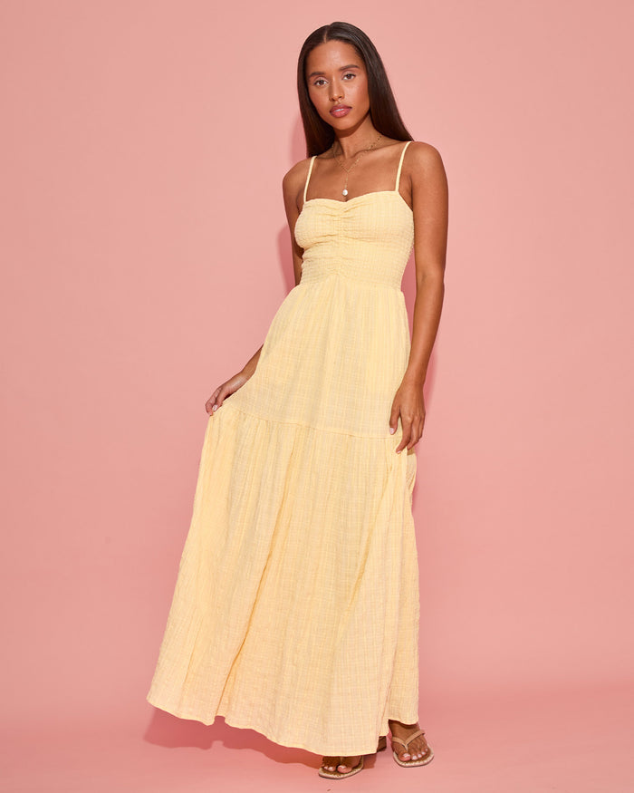 Front view of a model wearing the LSPACE Mallorca Dress in daisy, showing the smocked bodice and tiered maxi skirt in a soft yellow gauze fabric, photographed against a pink background.