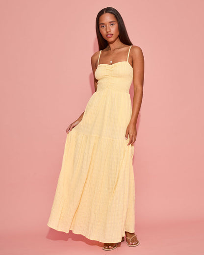 Front view of a model wearing the LSPACE Mallorca Dress in daisy, showing the smocked bodice and tiered maxi skirt in a soft yellow gauze fabric, photographed against a pink background.