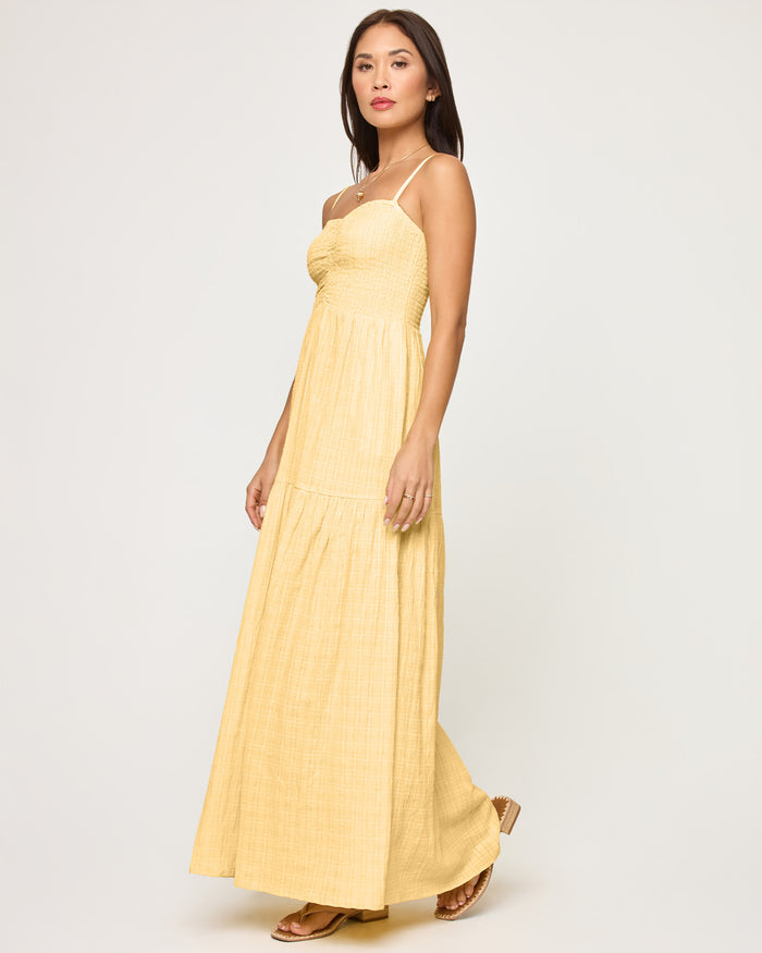 Side view of a model wearing the LSPACE Mallorca Dress in daisy, showing the smocked bodice and full tiered maxi skirt in soft yellow gauze fabric.
