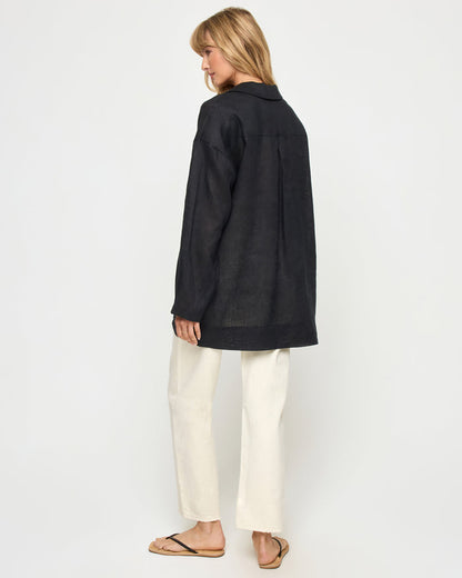 Back view of a model wearing the LSPACE Oasis Tunic in black, showing a collared neckline, long sleeves, and a relaxed oversized silhouette, styled with cream wide-leg pants.