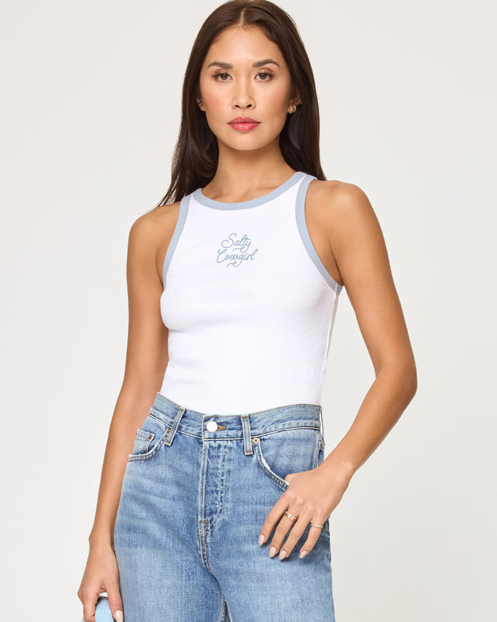 Front view of a model wearing the LSPACE Rewind Tank in salty cowgirl, a white ribbed tank top with blue contrast trim and a salty cowgirl script graphic on the chest, styled with high-waisted jeans.