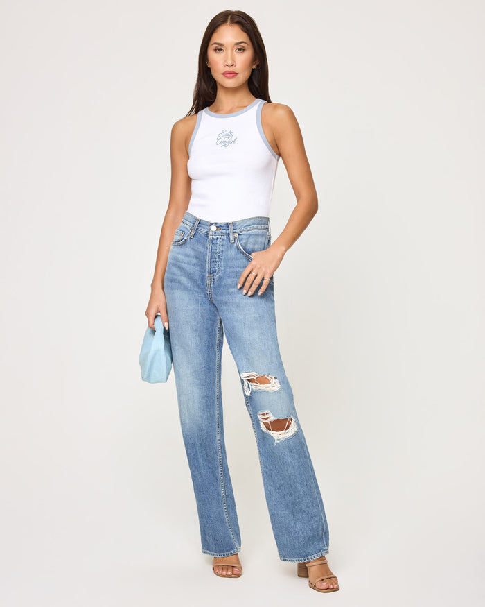 Front view of a model wearing the LSPACE Rewind Tank in salty cowgirl, a fitted white ribbed tank with blue contrast trim at the neckline and armholes and a salty cowgirl script graphic on the chest, styled with distressed wide-leg jeans.