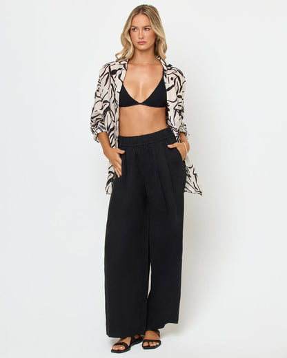 Front view of a model wearing the LSPACE Rio Tunic in heavy petal, an open-front linen button-up shirt with a cream and black bold botanical brushstroke print, styled over a black bikini top and wide-leg black pants.
