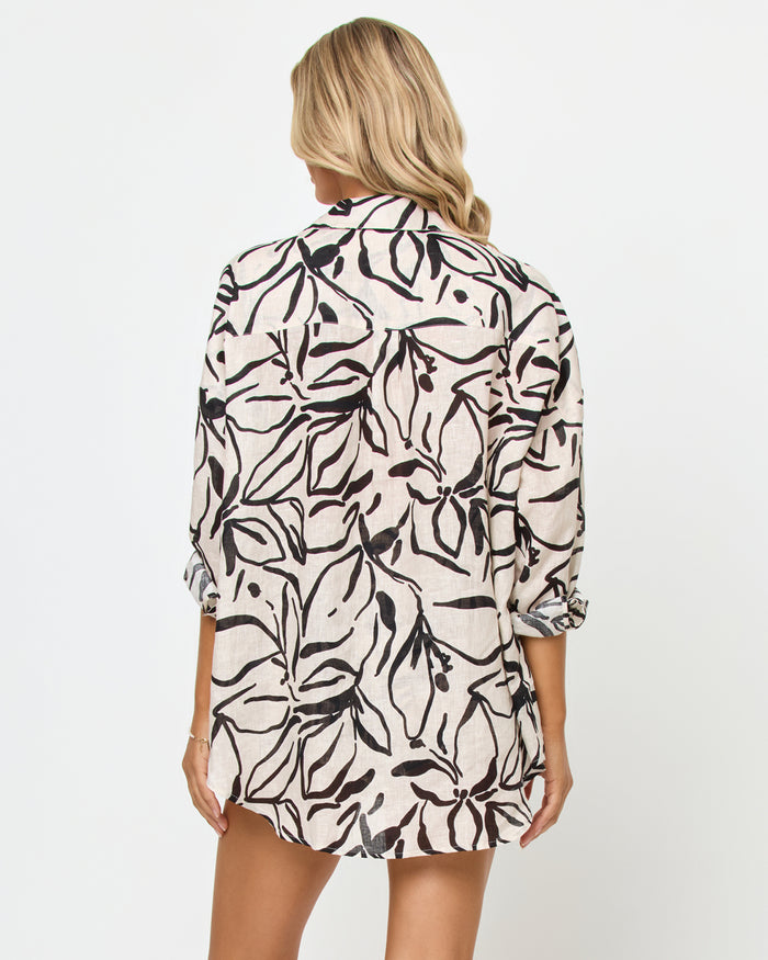 Back view of a model wearing the LSPACE Rio Tunic in heavy petal, showing the full back of the cream linen oversized button-up shirt with bold black botanical brushstroke print and three-quarter sleeves.
