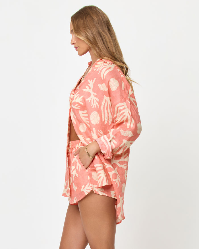 Side view of a model wearing the LSPACE Rio Tunic in shell or high water, showing the relaxed linen button-up silhouette with a coral and cream tropical seashell and coral print, styled with the matching Rio Short.