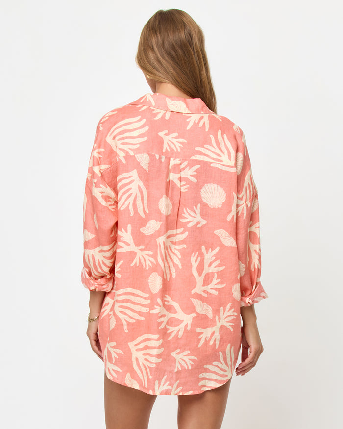 Back view of a model wearing the LSPACE Rio Tunic in shell or high water, showing the full back of the coral linen oversized button-up shirt with a cream tropical seashell and coral print.