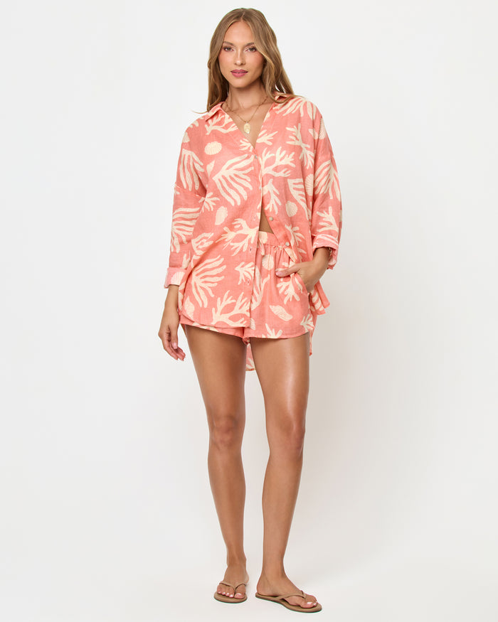 Front view of a model wearing the LSPACE Rio Tunic in shell or high water, a coral linen button-up shirt with a cream tropical seashell and coral print and three-quarter sleeves, styled with the matching Rio Short and tan sandals.