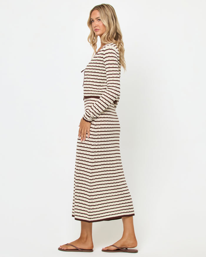 Side view of a model wearing the LSPACE Rosalie Cardigan in cafe-cream, a cropped knit cardigan with cream and dark brown horizontal stripes, styled with the matching Rosalie Maxi dress and brown sandals.