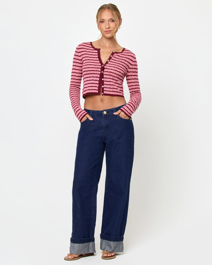 Front view of a model wearing the LSPACE Rosalie Cardigan in strawberry stripe, a cropped knit cardigan with pink and burgundy horizontal stripes, a V-neckline, and gold button closure, styled with wide-leg dark wash jeans and sandals.