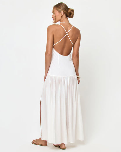 Back view of a model wearing the LSPACE Row Maxi Dress in cream, a white sleeveless maxi dress with criss-cross spaghetti straps, a low open back, and a flowy pleated skirt with a side slit, styled with tan sandals and a gold bangle.