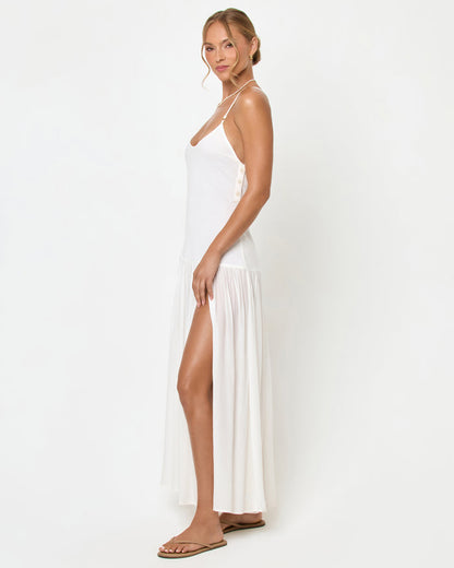 Side view of a model wearing the LSPACE Row Maxi Dress in cream, a white sleeveless maxi dress with thin spaghetti straps, a fitted bodice, and a flowy pleated skirt with a high side slit, styled with tan sandals.