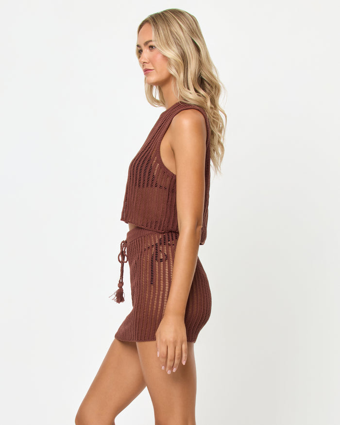 Side view of a model wearing the LSPACE Sander Skirt in cafe, a crochet mini skirt with a tassel drawstring waist, styled with the matching crochet crop top.