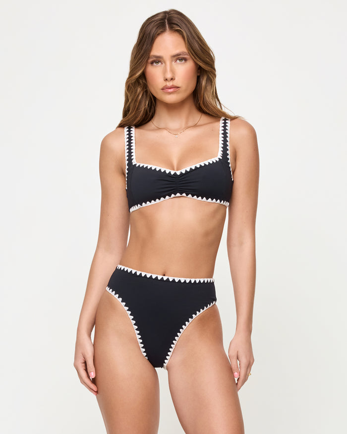 Front view of a model wearing the LSPACE Black-Cream Savannah Bikini Bottom, a high-waist black bottom with cream zigzag crochet trim, paired with a matching black square-neck bralette Effie Bikini Top with cream zigzag trim.