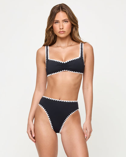 Front view of a model wearing the LSPACE Black-Cream Savannah Bikini Bottom, a high-waist black bottom with cream zigzag crochet trim, paired with a matching black square-neck bralette Effie Bikini Top with cream zigzag trim.