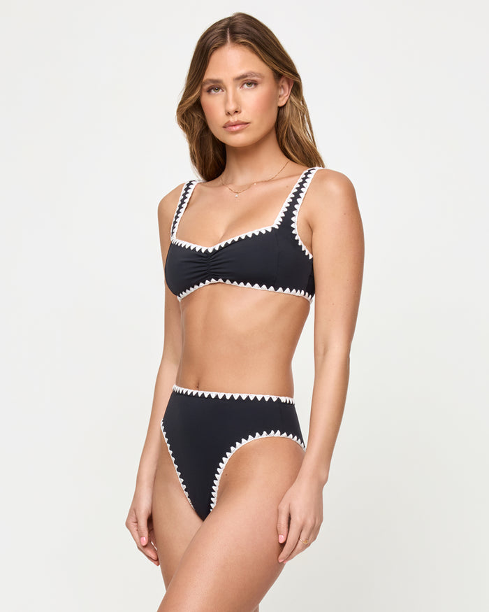 Three-quarter front view of a model wearing the LSPACE Black-Cream Savannah Bikini Bottom with cream zigzag crochet trim, styled with a matching black bralette Effie Bikini Top with cream zigzag trim.