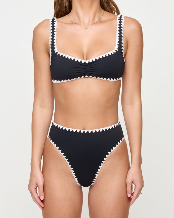 Cropped front close-up of a model wearing the LSPACE Black-Cream Savannah Bikini Bottom, highlighting the cream zigzag crochet trim detail on the waistband and leg openings, styled with a matching black bralette Effie Bikini Top.