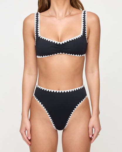 Cropped front close-up of a model wearing the LSPACE Black-Cream Savannah Bikini Bottom, highlighting the cream zigzag crochet trim detail on the waistband and leg openings, styled with a matching black bralette Effie Bikini Top.