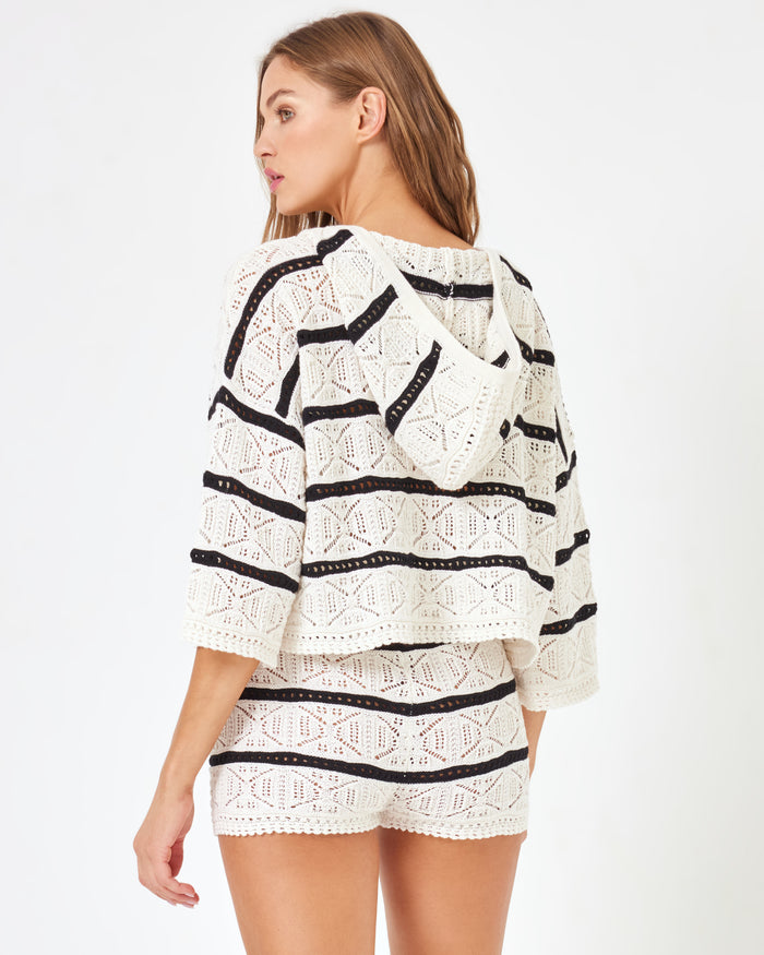 Back view of a model wearing the LSPACE Striped Diamond Eyes Short in Starcrossed Stripe, cream open-knit crochet shorts with black horizontal stripe trim, styled as a matching set with the coordinating crochet hoodie.
