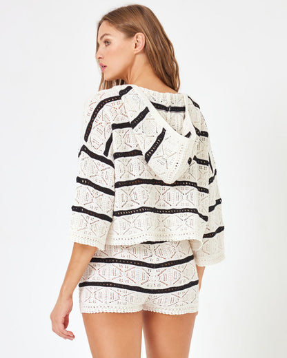 Back view of a model wearing the LSPACE Striped Diamond Eyes Short in Starcrossed Stripe, cream open-knit crochet shorts with black horizontal stripe trim, styled as a matching set with the coordinating crochet hoodie.