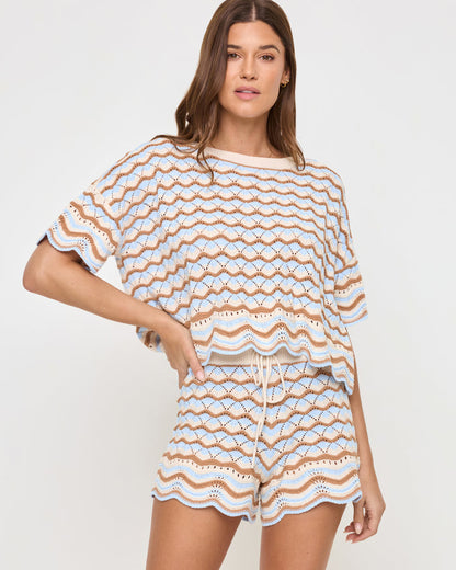 Front view of a model wearing the LSPACE Sunchaser Short in north shores, a crochet short with a drawstring waist and a wave scallop pattern in light blue and tan, paired with the coordinating knit top.